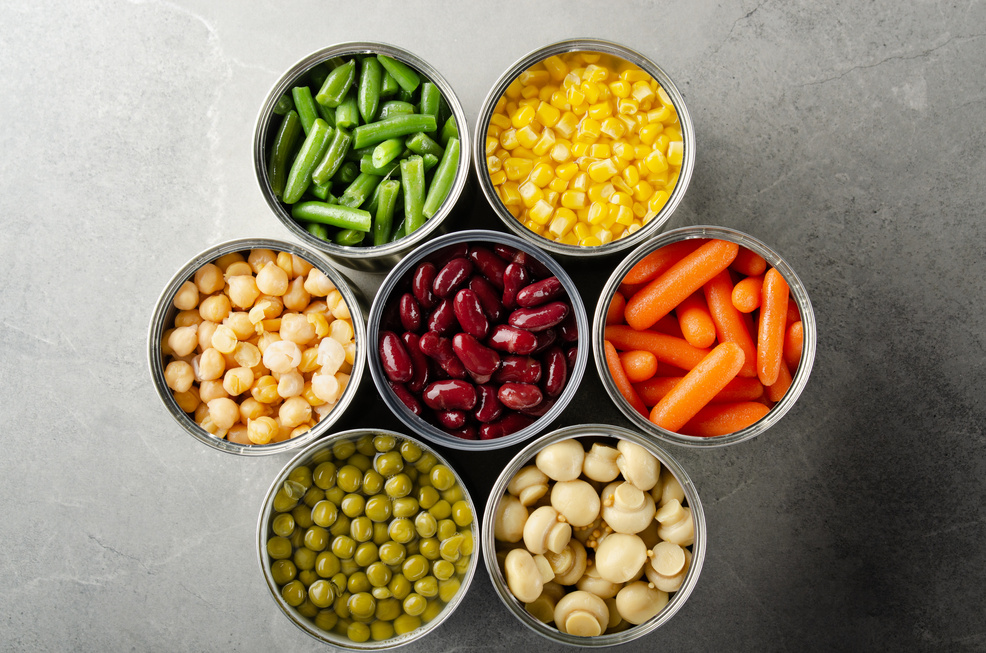 Flat lay view at canned vegetables in opened tin cans on kitchen table. Non-perishable long shelf life foods background